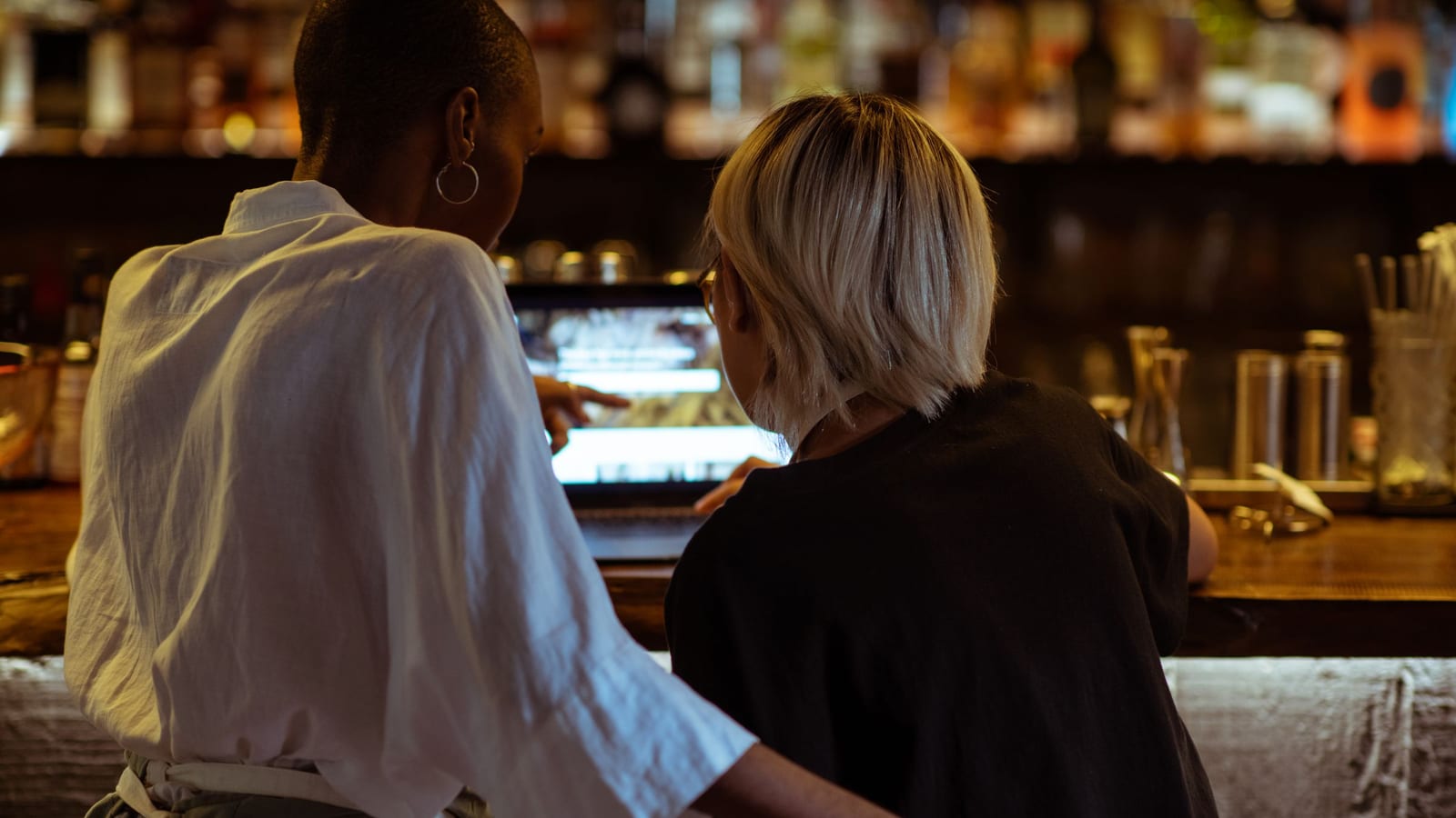 Two restaurant workers, seen from behind, look over a notebook computer together at their bar counter before service, planning the week’s Facebook ads.