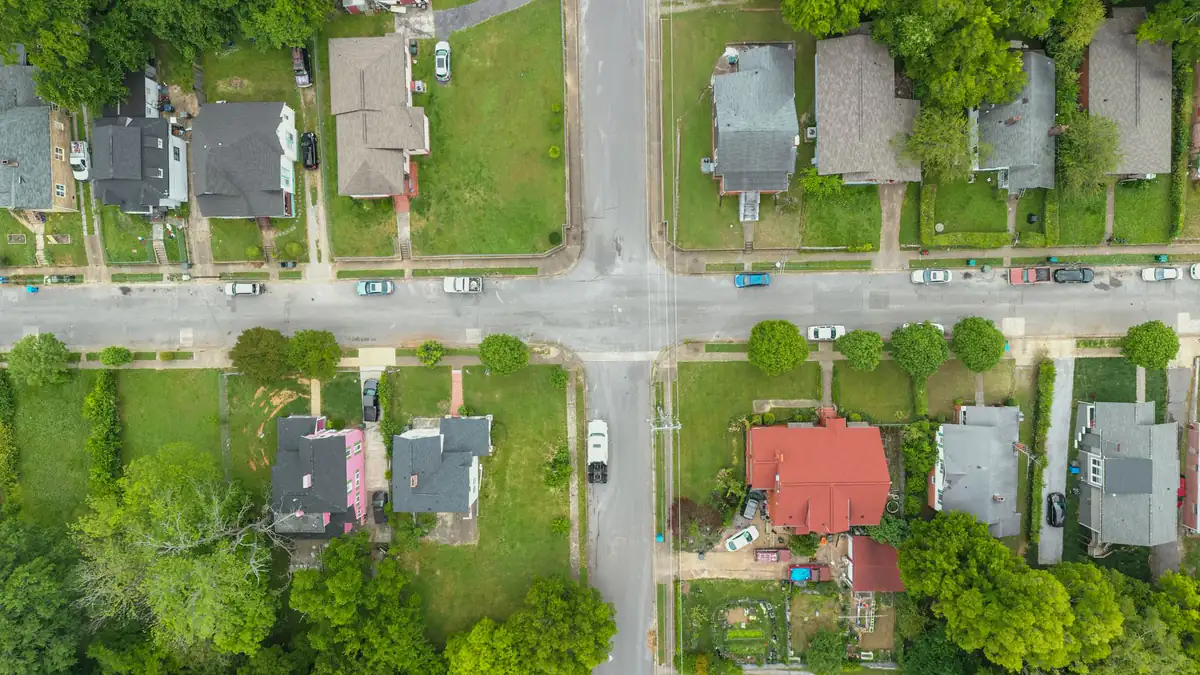Seen from straight above, two streets cross between rows of houses, lawns, parked cars and trees.