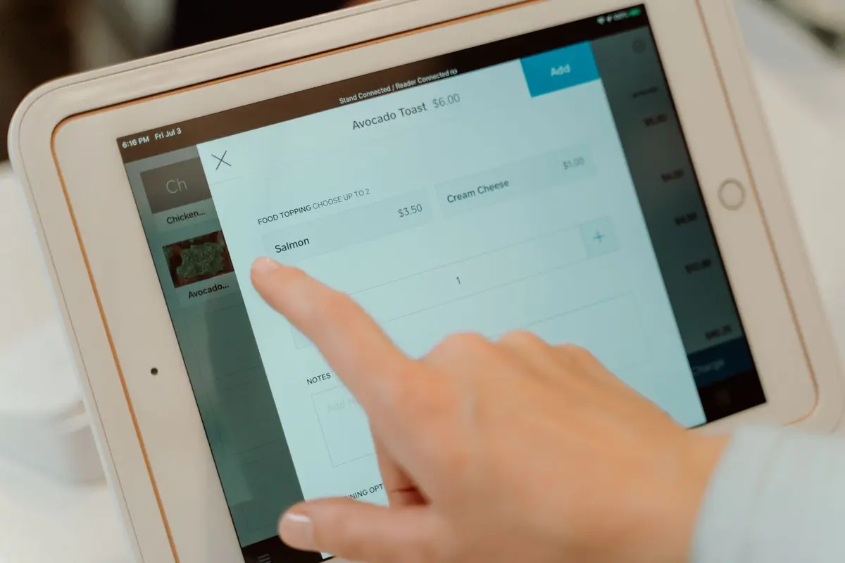 A finger taps a menu item on a tablet register at a counter.