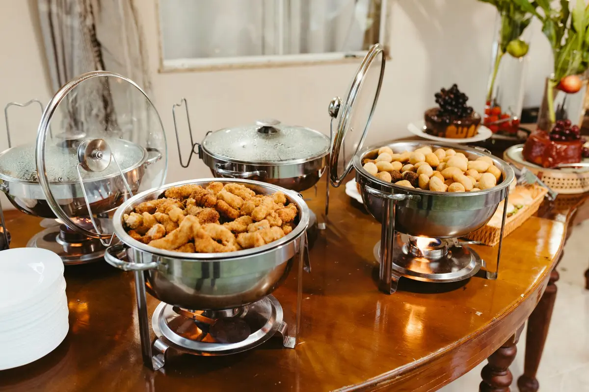 Round chafing dishes over lit fuel burners on a wooden party table, one full of breaded fried bites, with a stack of plates beside them.