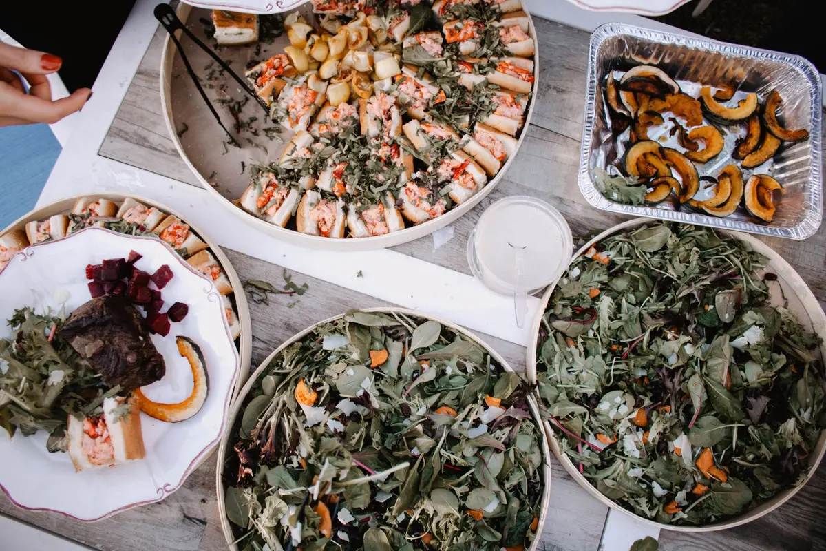 Party trays seen from above: platters of mini sandwich rolls, two big green salads and a foil pan of roasted squash, with one hand reaching in.