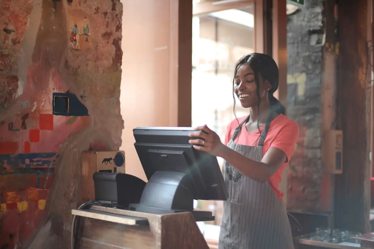A smiling cashier in a striped apron rings up an order on a counter screen in a brick-walled café.