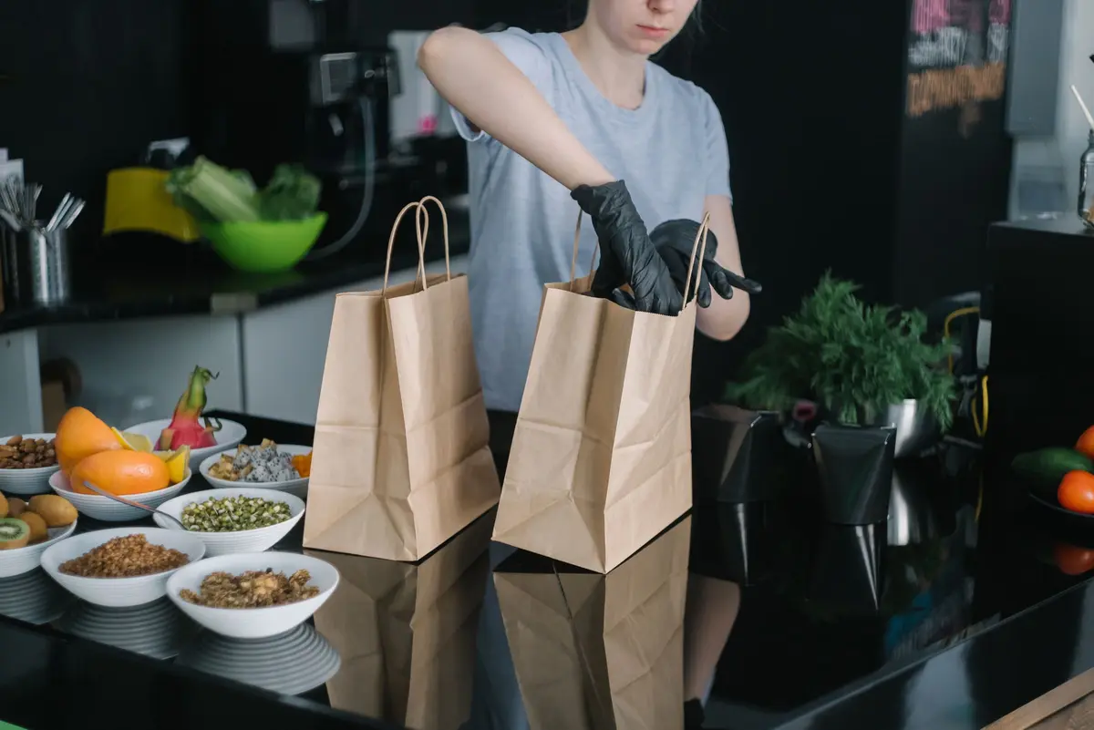 A worker in black gloves packs two kraft paper takeout bags on a dark counter beside bowls of food.