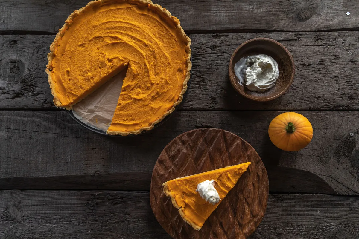 A pumpkin pie with one slice cut out, the slice on a wooden board beside a bowl of whipped cream.