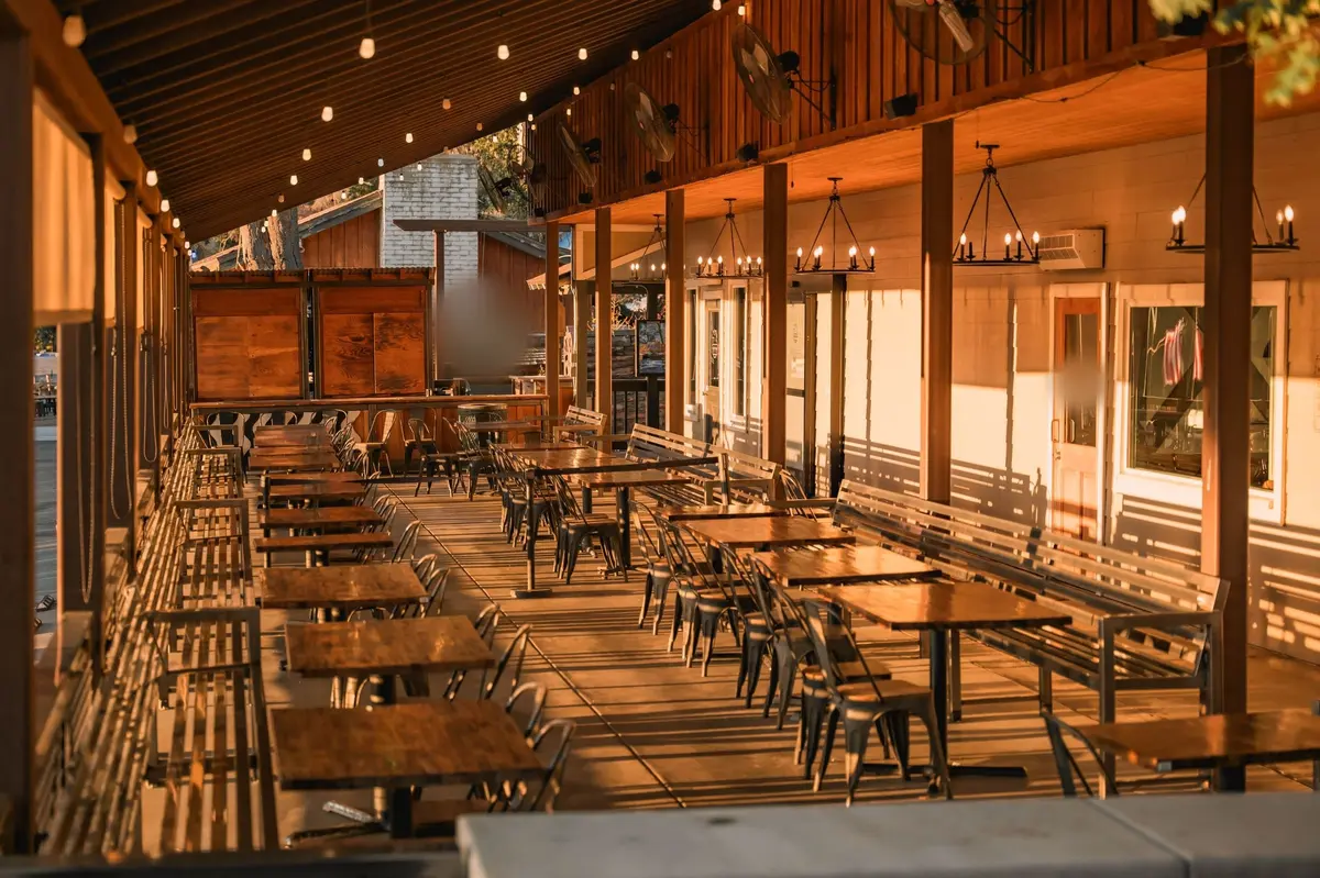 An empty covered restaurant patio with rows of wooden tables, string lights and candle chandeliers, lit by low evening sun.