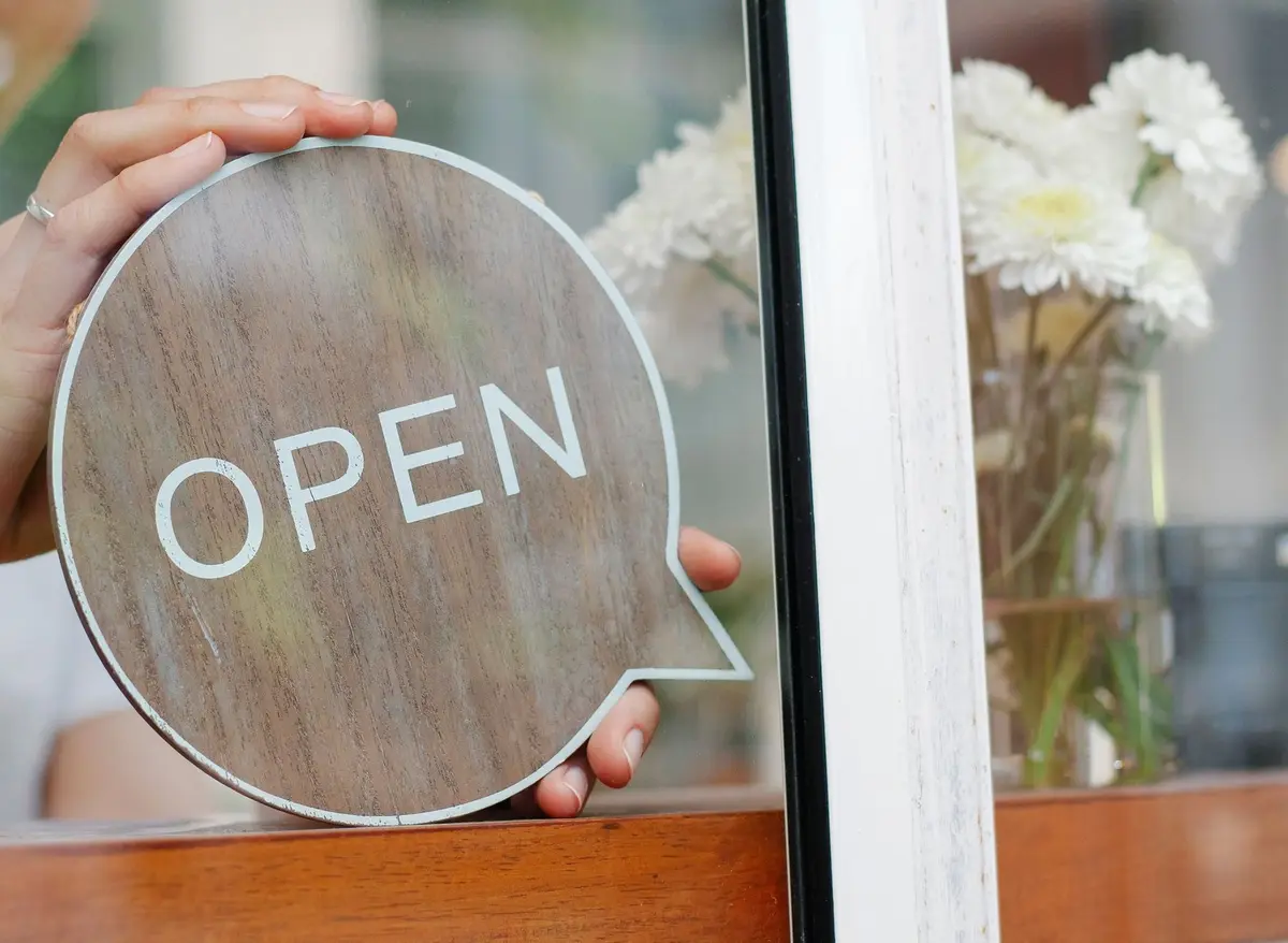 A staff member’s hands set a round wooden OPEN sign against the glass of a shop window at opening time.