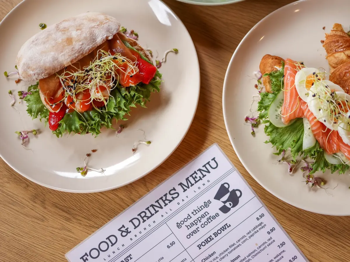 A printed food and drinks menu with prices lies on a wooden table beside two plated sandwiches.