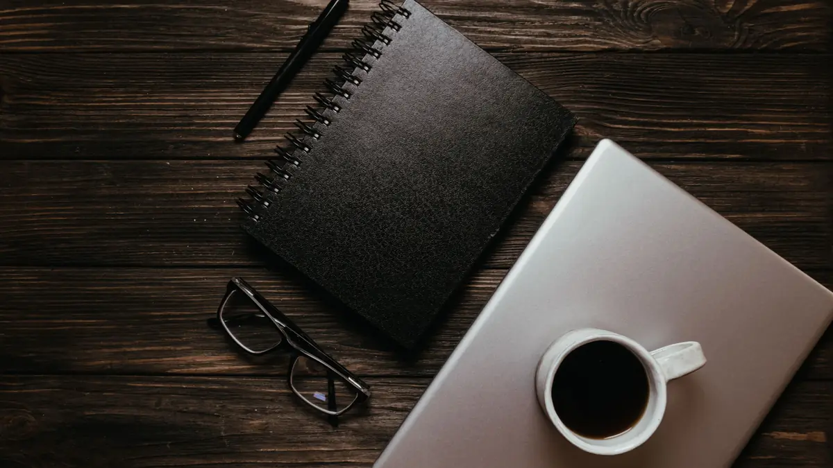 A closed laptop with a cup of coffee on it, beside a notebook, a pen and glasses on a dark wooden table.