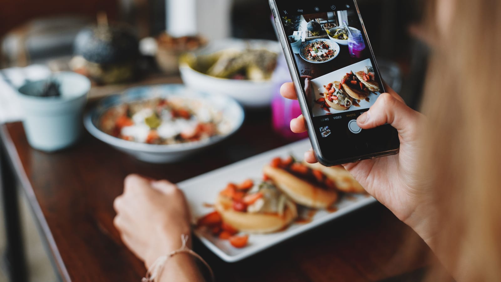 A diner photographs a plate of food on a phone at a cafe table, with more dishes in front of her.