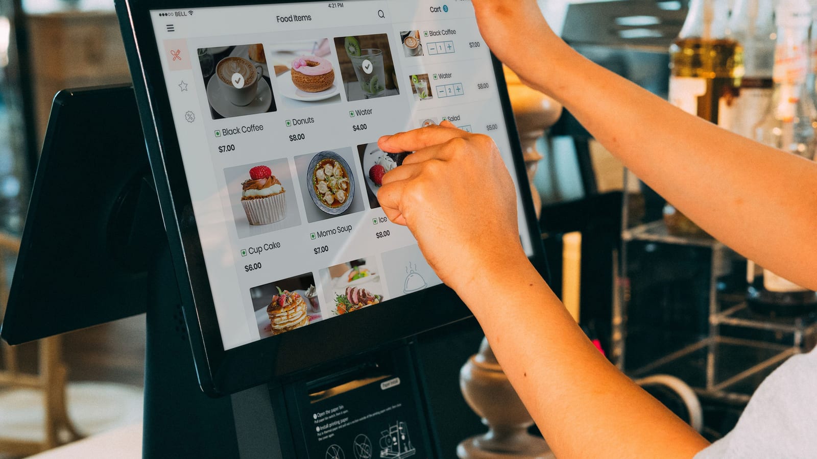 A restaurant worker’s hand taps a dish on a counter touchscreen, where sales results are recorded item by item