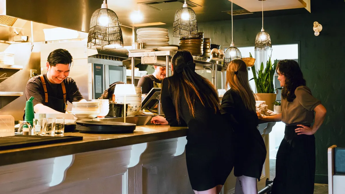 A smiling cook works an open kitchen under pendant lamps while three front-of-house staff talk at the counter, one folding napkins
