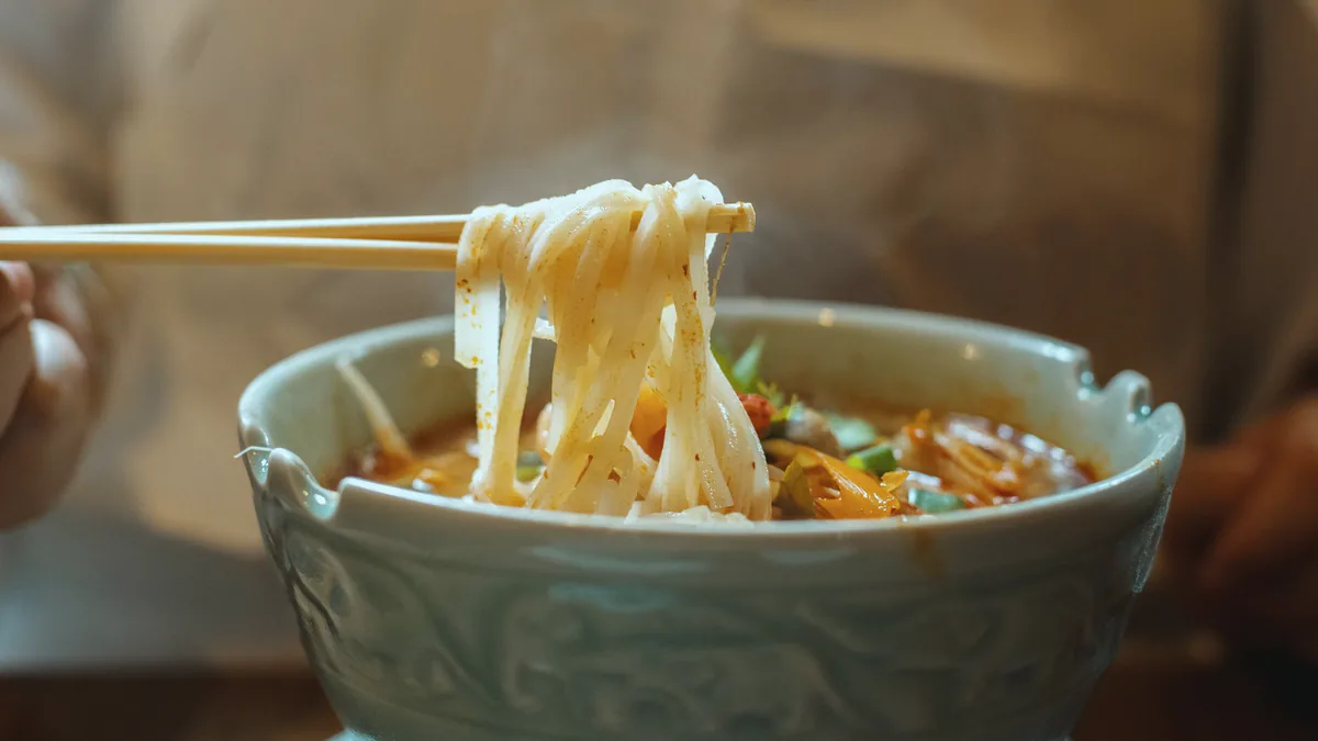 Chopsticks lift wide noodles from a pale green bowl of noodle soup, the room behind it soft and warm