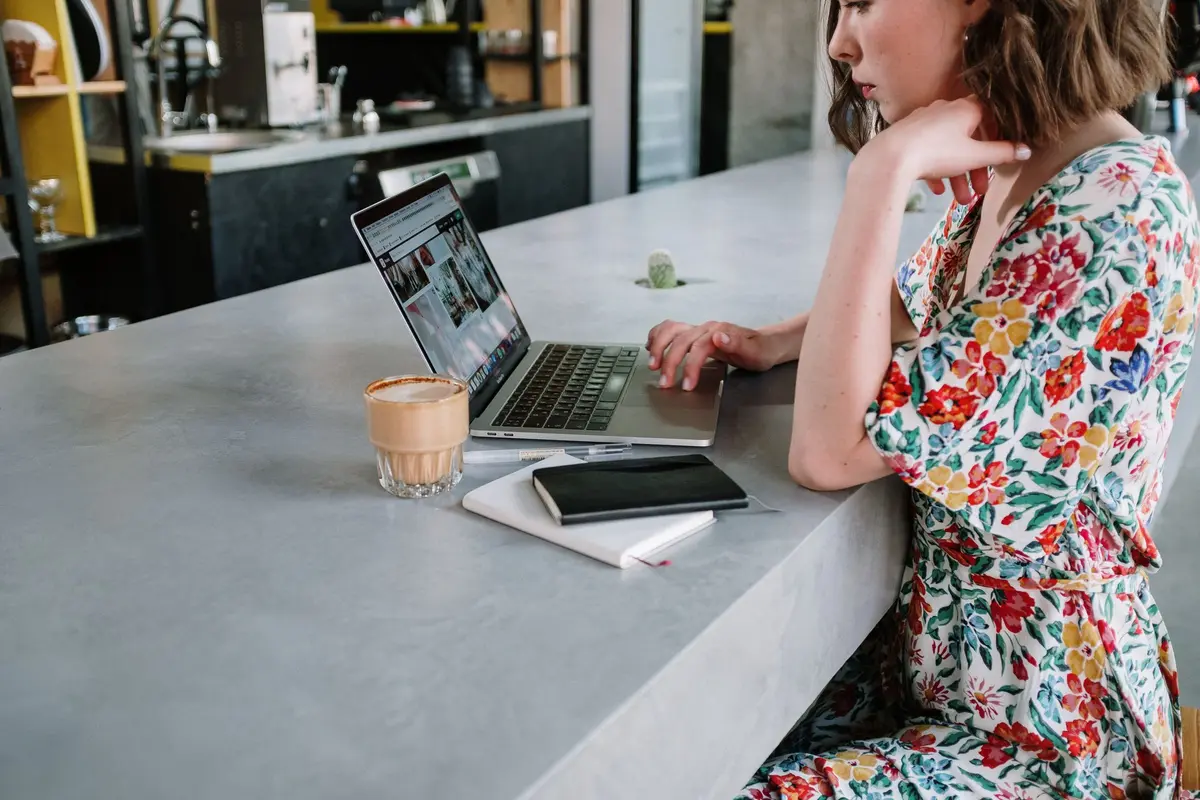 A woman works on a laptop at a cafe counter, a coffee and a notebook beside her.