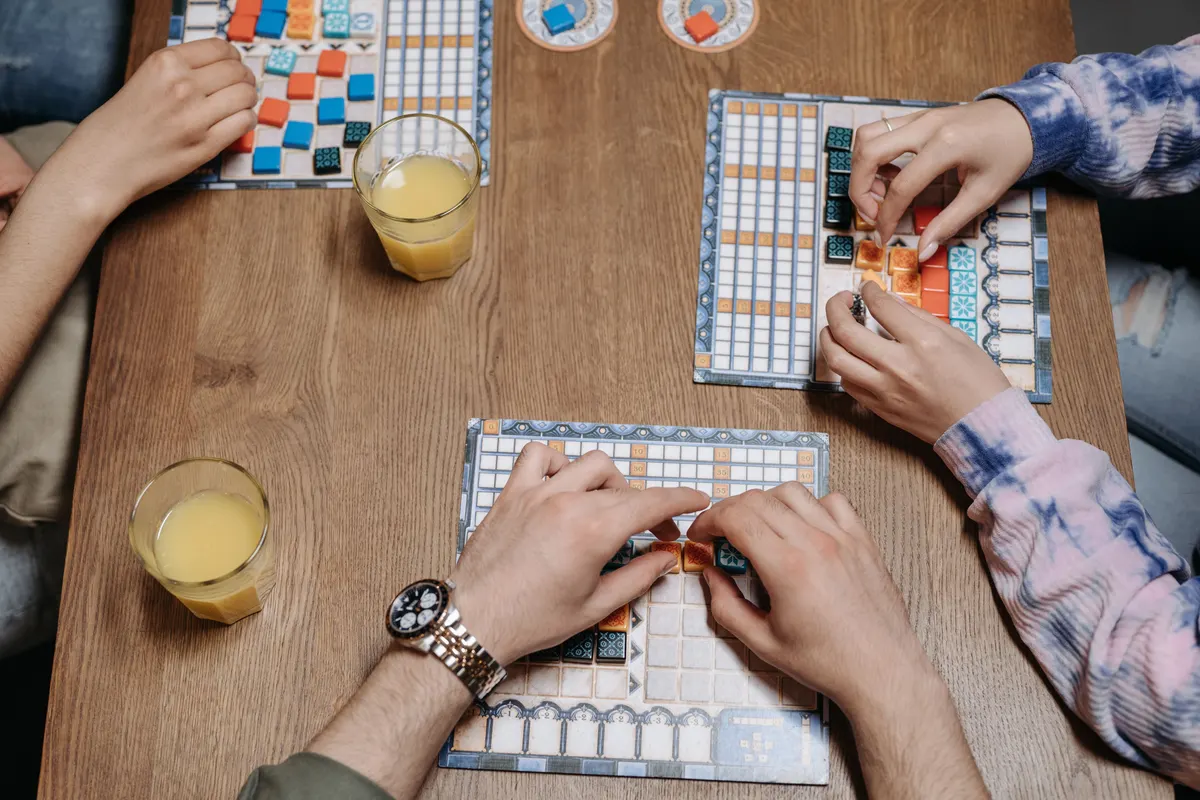 Seen from above, guests place tiles on board games at a wooden restaurant table, glasses of juice beside them.