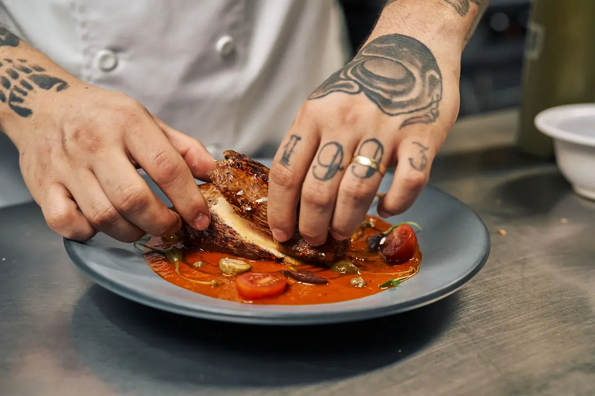 A cook’s tattooed hands press a seared chicken thigh into a bright tomato sauce on a gray plate.