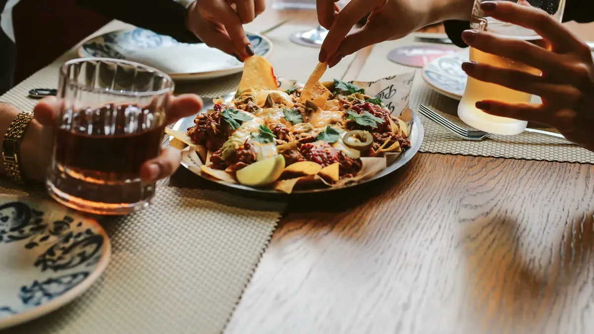 Two friends reach into a plate of nachos on a wooden restaurant table, a soda and a beer in their other hands