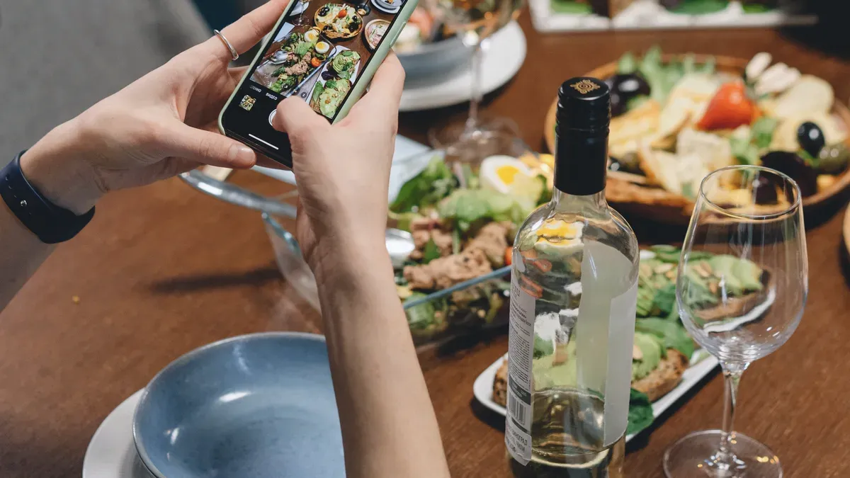 Two hands hold a phone over a wooden table of shared plates and wine glasses, the dishes showing on the phone screen