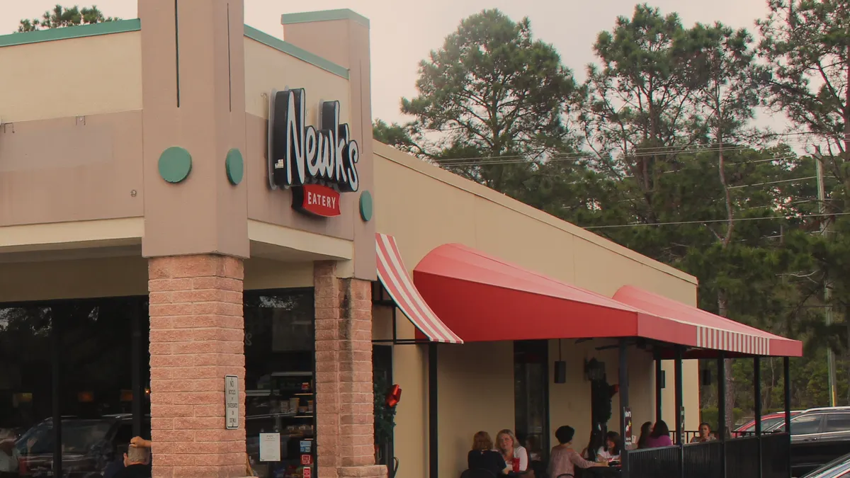 A Newk's Eatery sign on the corner tower of a strip-center store, a red awning over the patio beside it.