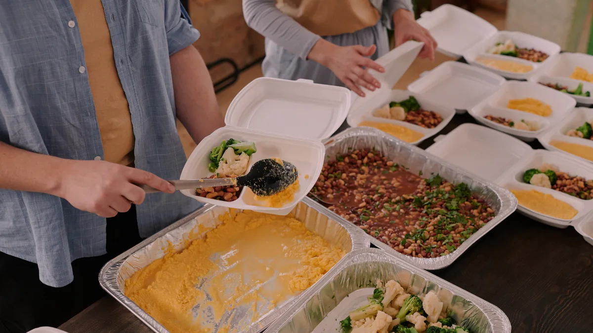 Hands spoon food into foam boxes beside foil trays of grains, a green salad and a baked dish, on a long table of open boxes.