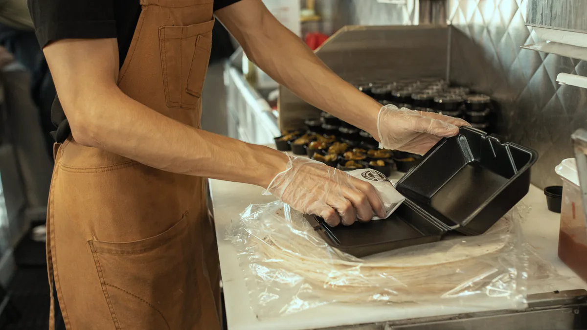 A cook in a brown apron and plastic gloves closes a black takeout container at a prep counter, more containers stacked behind.