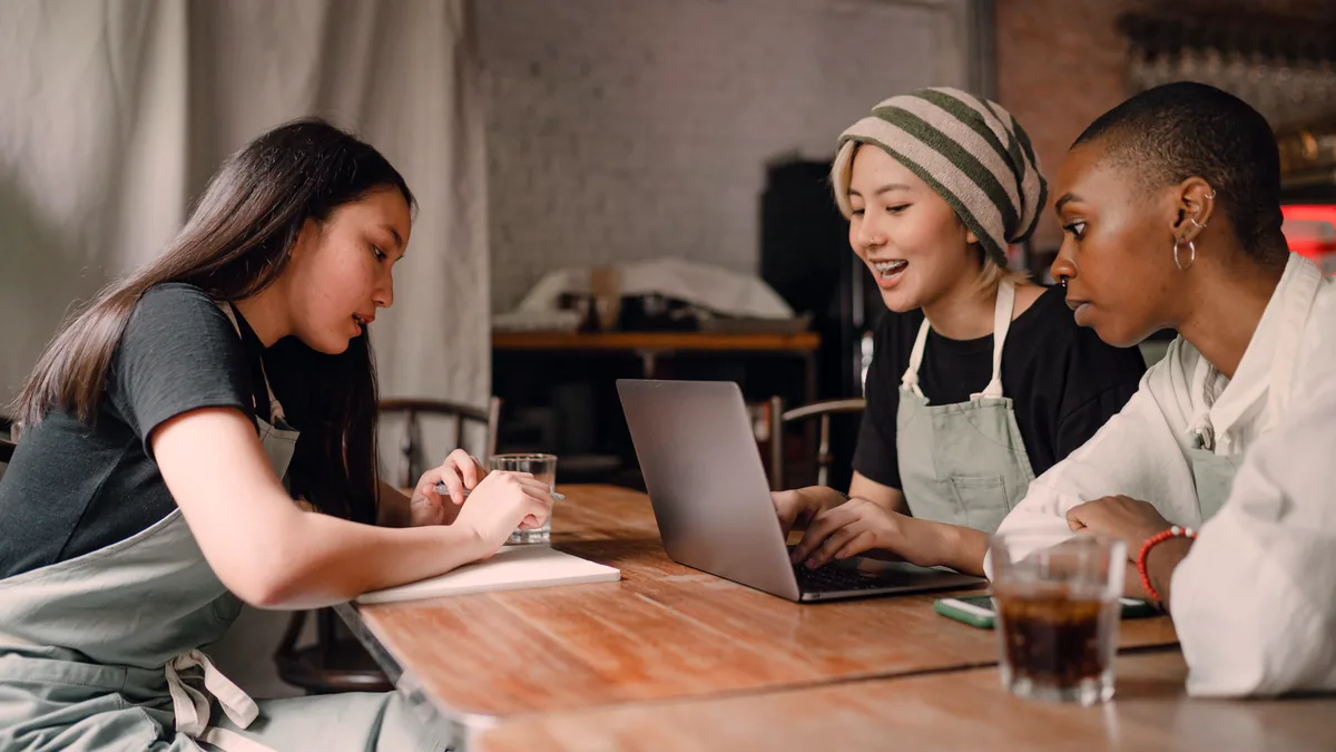 Three cafe workers in aprons sit at a wooden table around a laptop, one taking notes while the other two read the screen.
