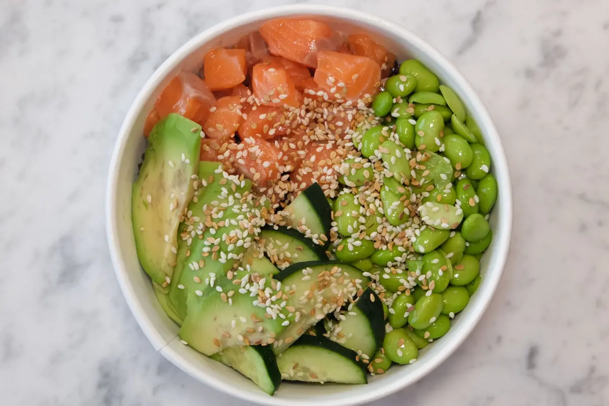 A poke bowl seen from above, with raw salmon, avocado, cucumber, edamame and sesame seeds.