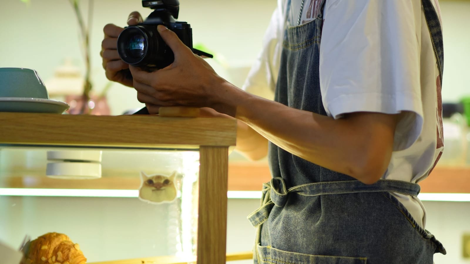 A café worker in a denim apron aims a small camera at a cup on the counter, the kind of photo a restaurant posts and may later boost. The face is out of frame.