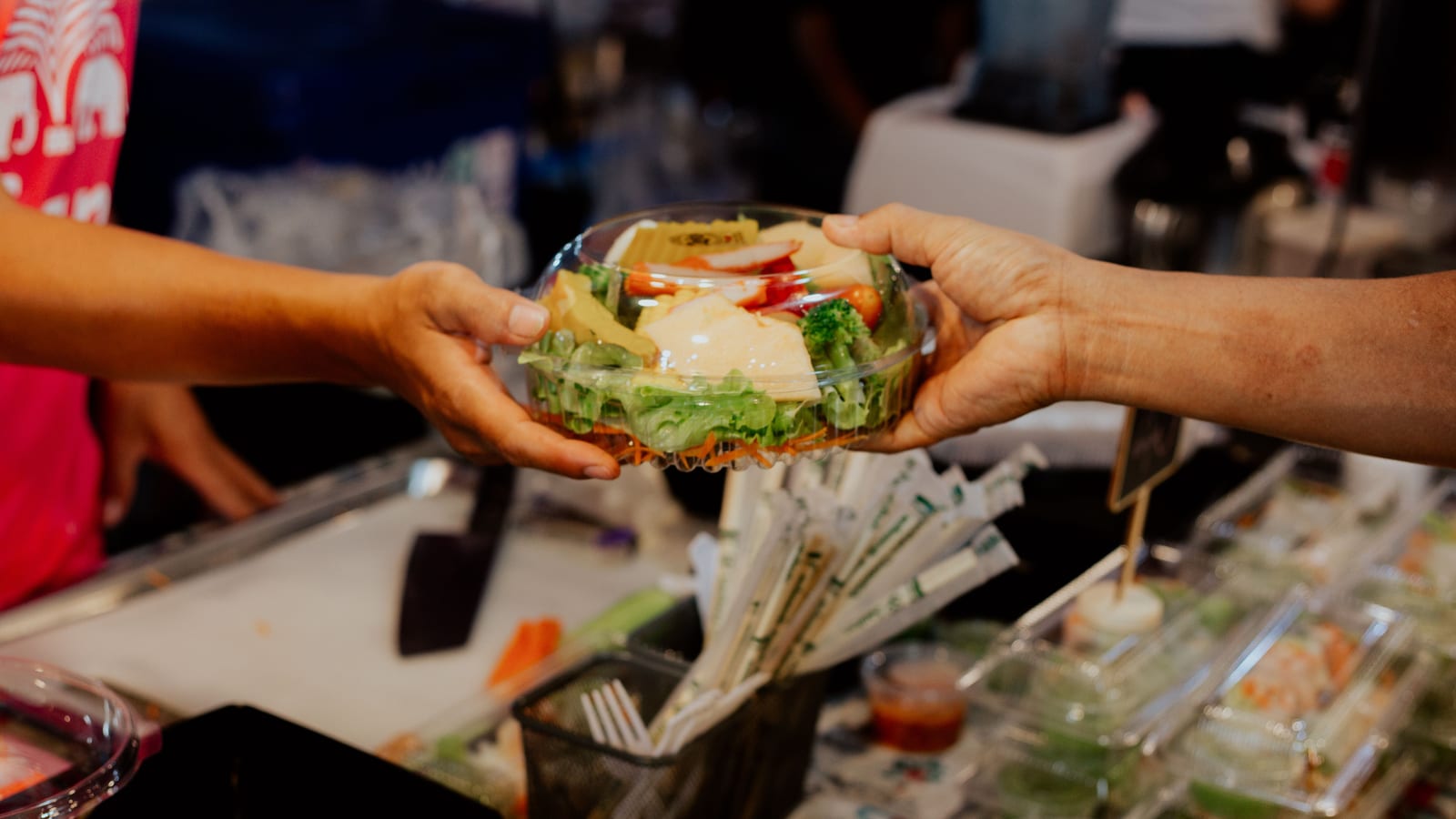 Across a restaurant counter, a worker hands a packed salad bowl to a guest.