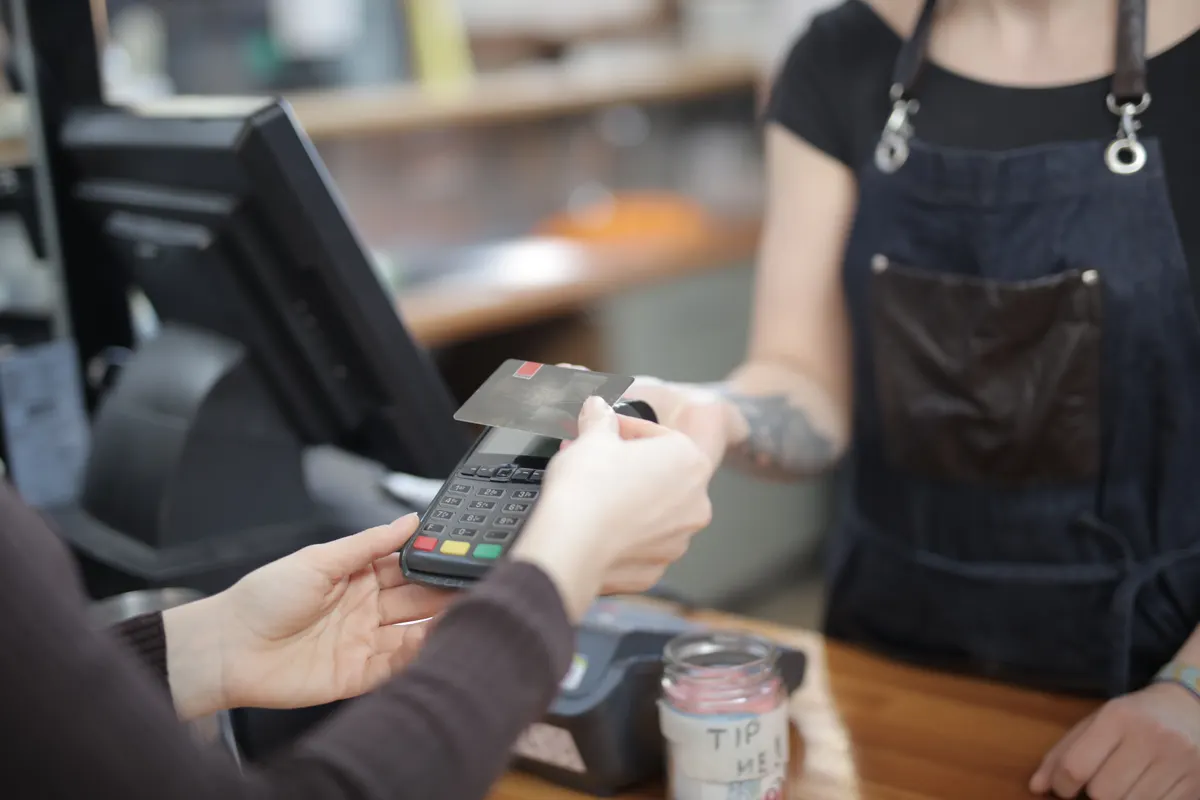 A guest holds a card to the payment terminal a server holds out across a café counter.