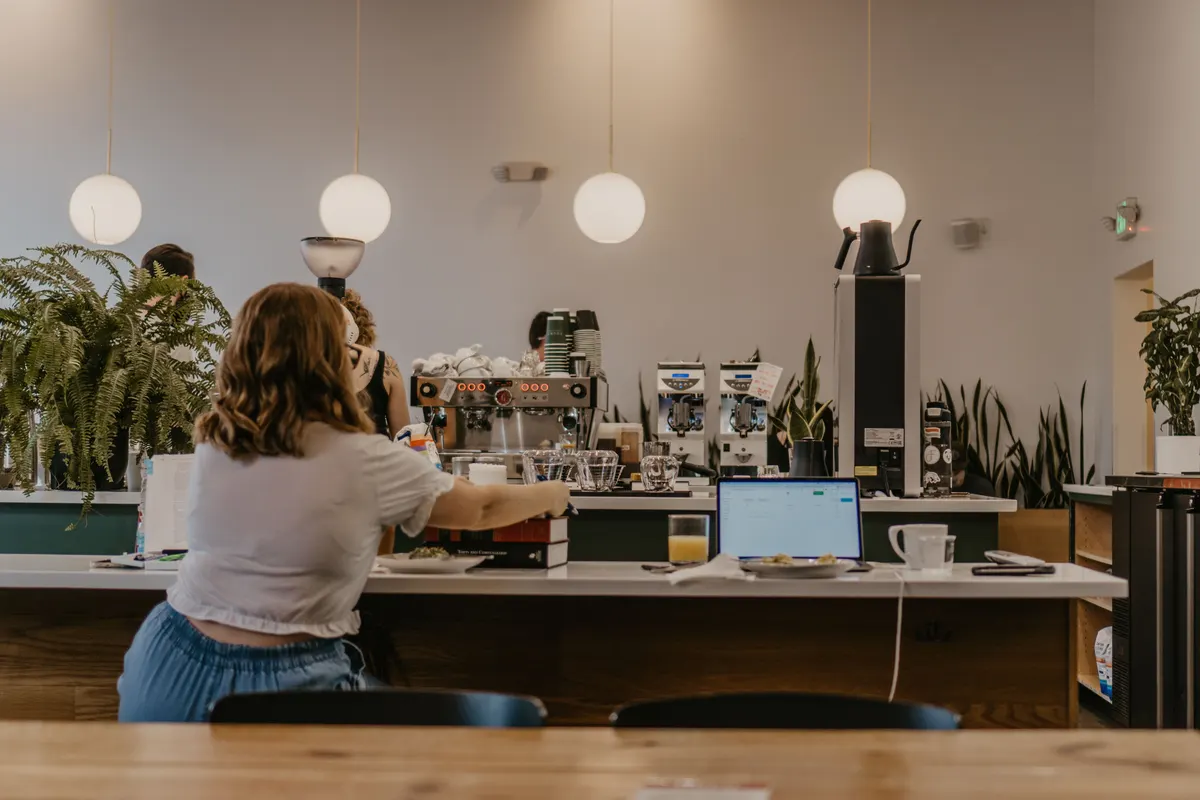 A guest sits alone at the long counter of a bright café, the espresso bar beyond.