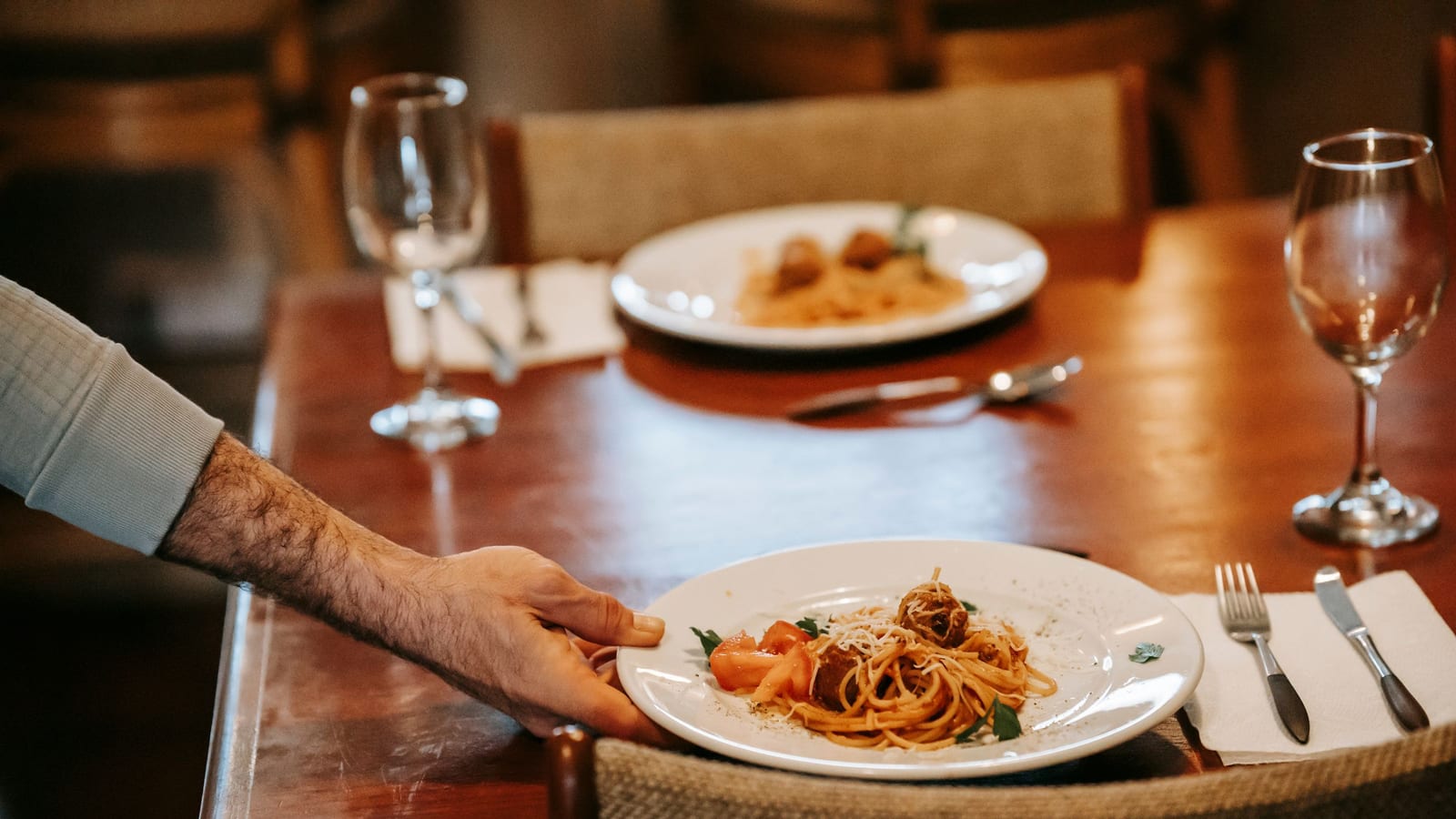 A server’s hand sets a plate of spaghetti and meatballs on a table for two, the kind of free meal a guest should still tip on.