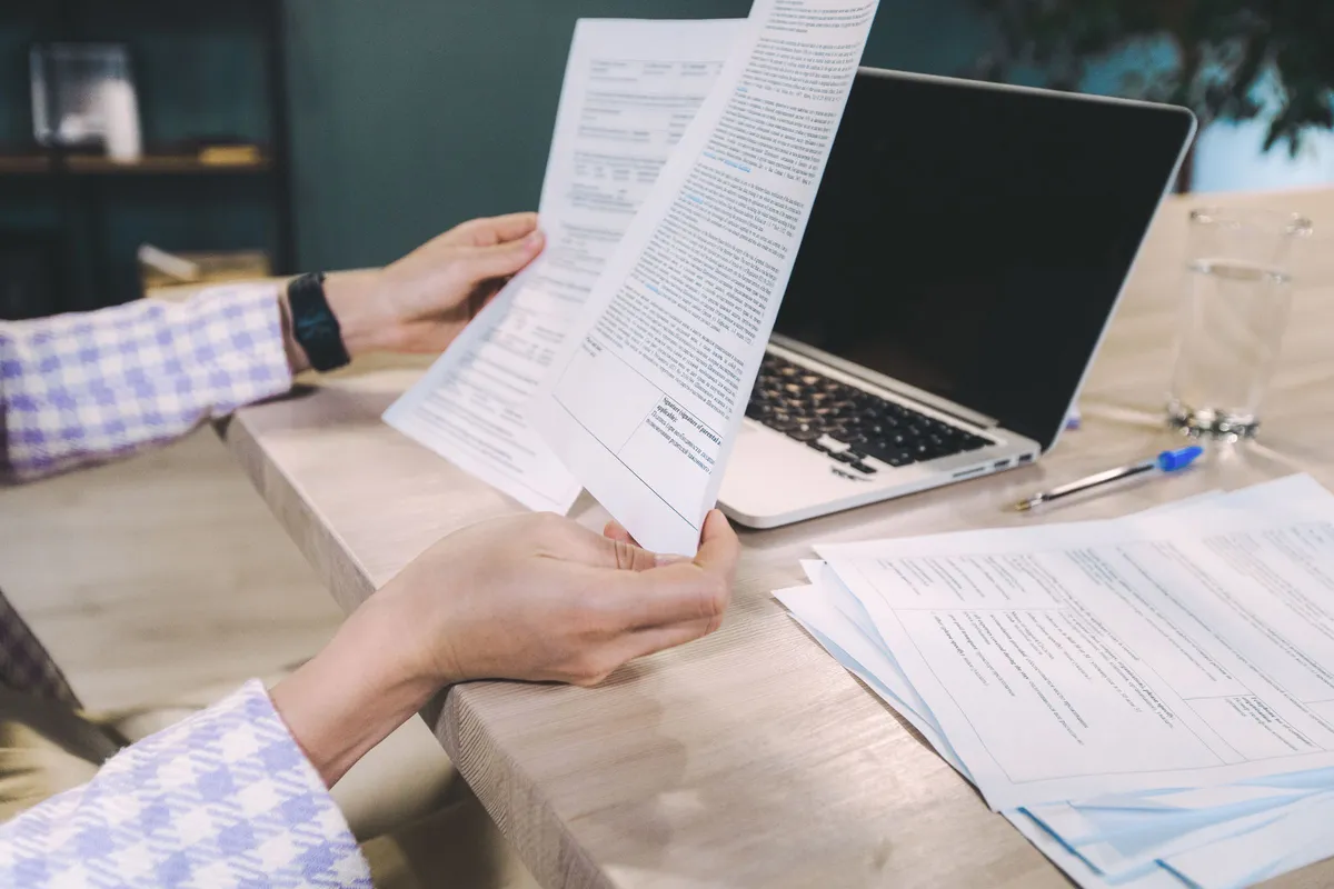 Hands hold printed pages beside an open laptop and a stack of papers on a wooden desk.