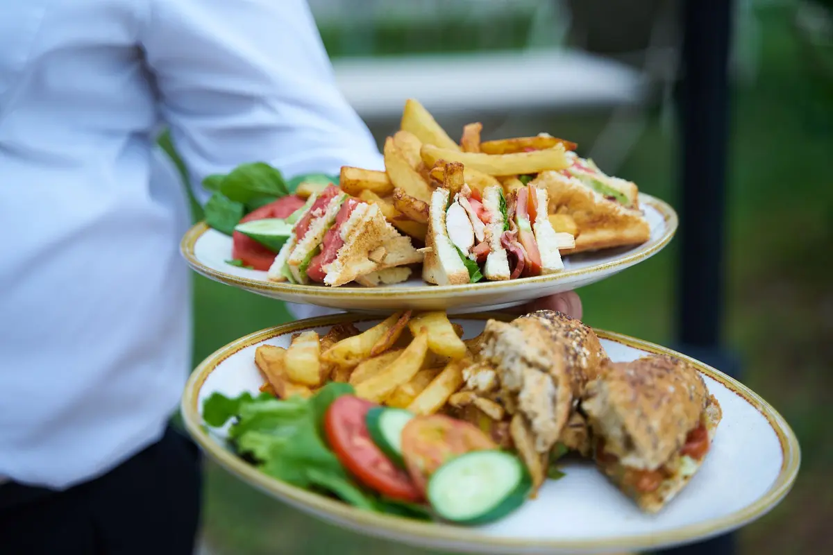 A server in a white shirt carries two plates of sandwiches and fries, face out of frame.