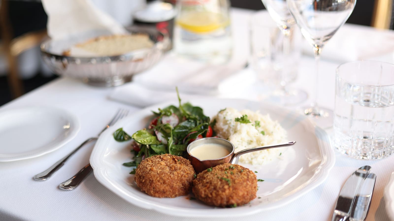 A restaurant plate on a white tablecloth: two breaded croquettes, rice, a green salad and a small pot of sauce.
