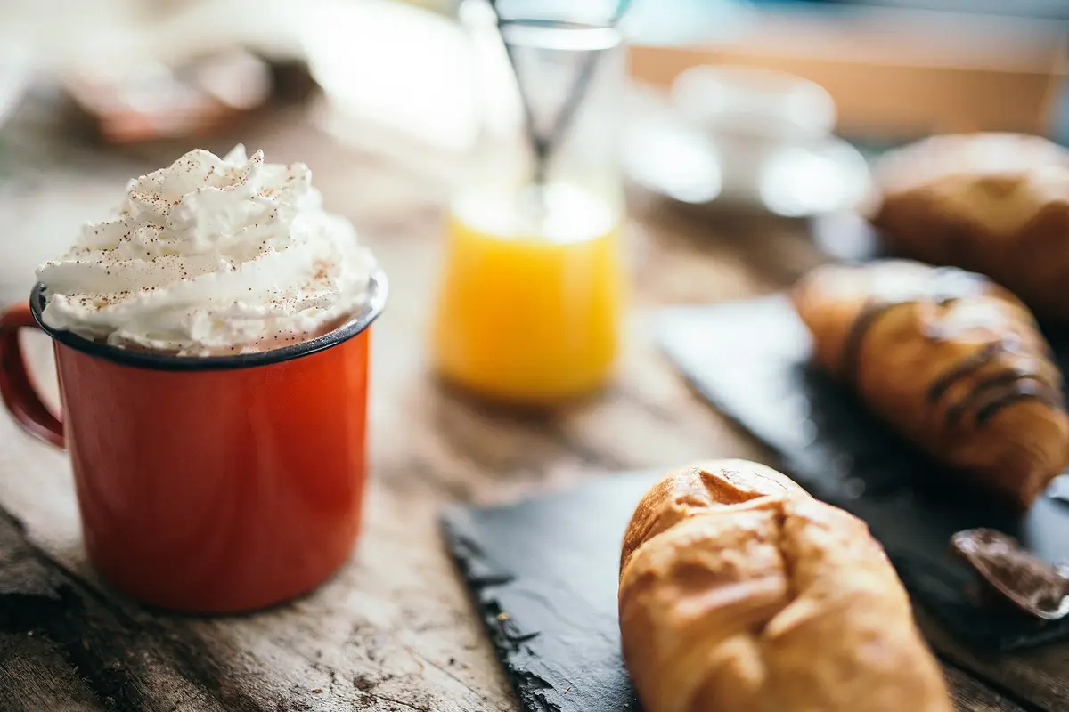 A red mug of hot drink topped with whipped cream sits beside croissants on a wooden café table.