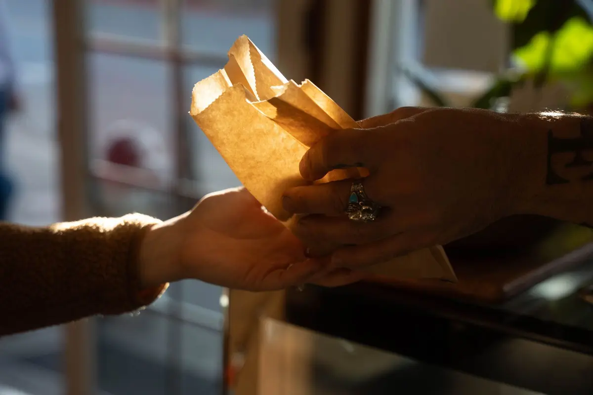 A staff member’s hand passes a paper pastry bag across a café counter into a guest’s open hand.