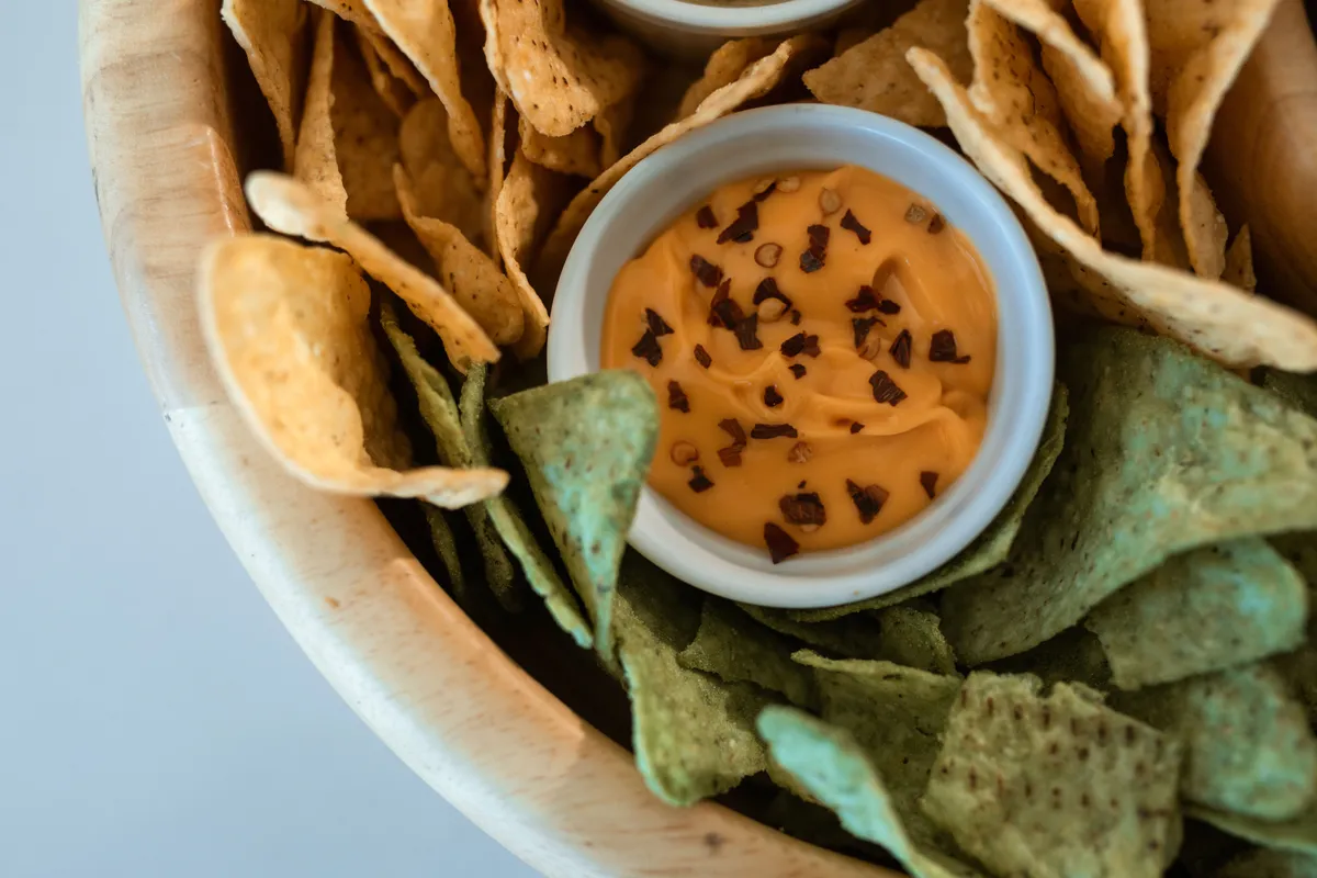 A small white cup of orange cheese dip with chile flakes sits in a wooden bowl of yellow and green tortilla chips.