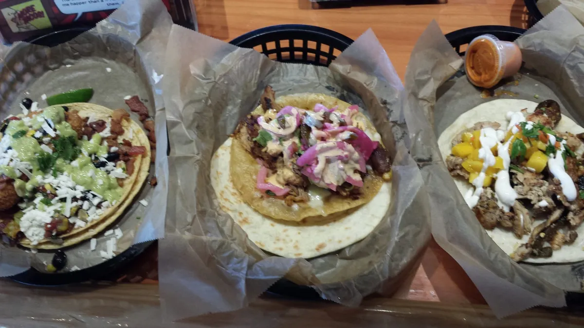 Three Torchy’s tacos sit in black baskets lined with paper on a wooden table in Waco, Texas, one with a sauce cup.