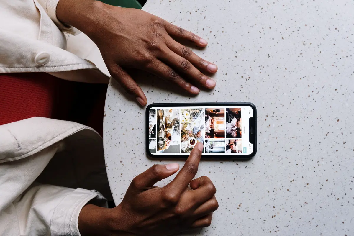 Two hands rest on a speckled cafe table beside a phone showing a grid of food photos.
