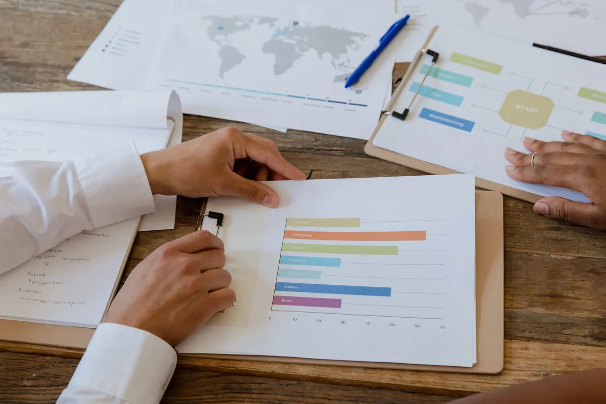 Hands rest on printed bar charts and a world map spread across a wooden table.