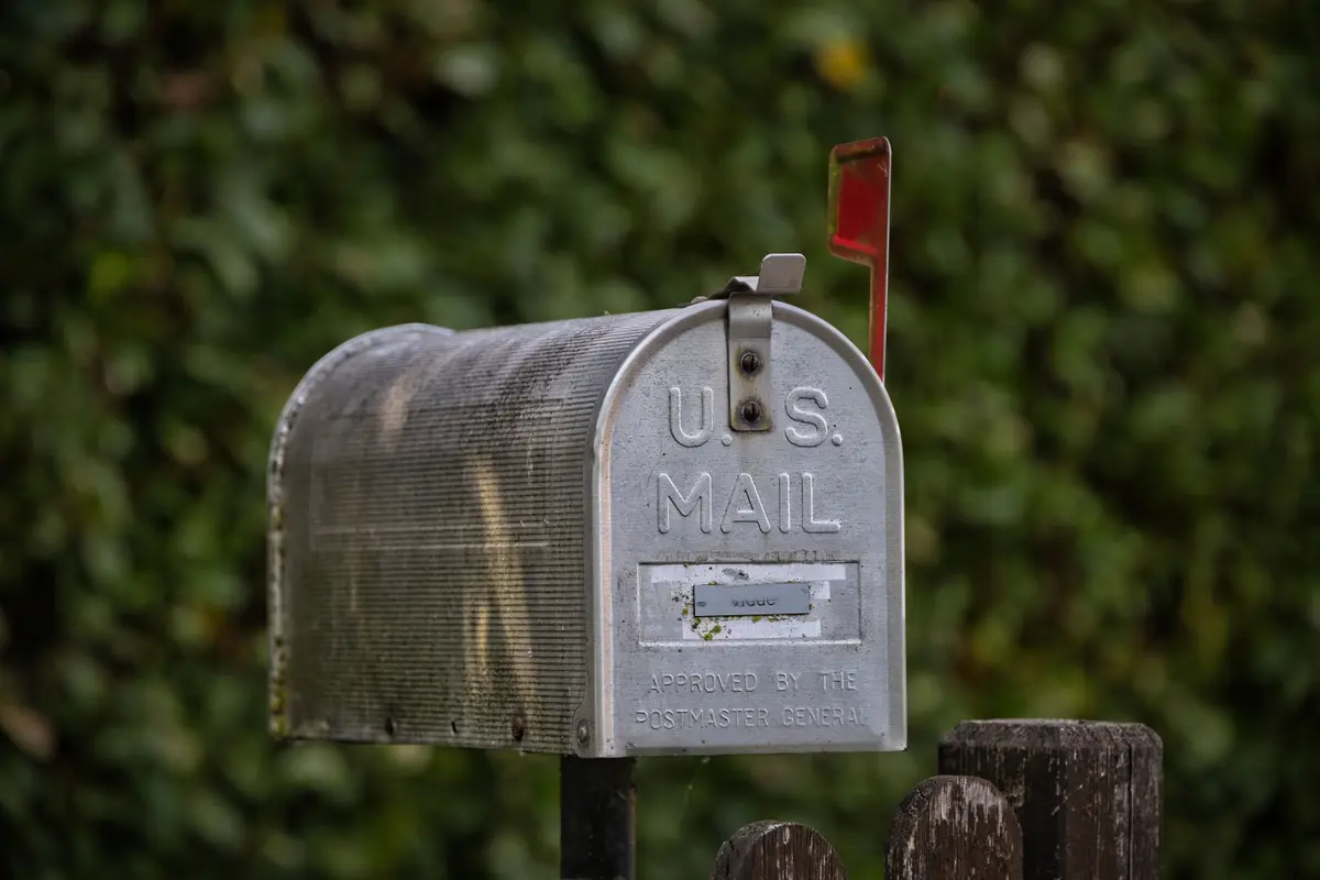 A silver curbside mailbox marked U.S. Mail, its red flag up, in front of a green hedge.