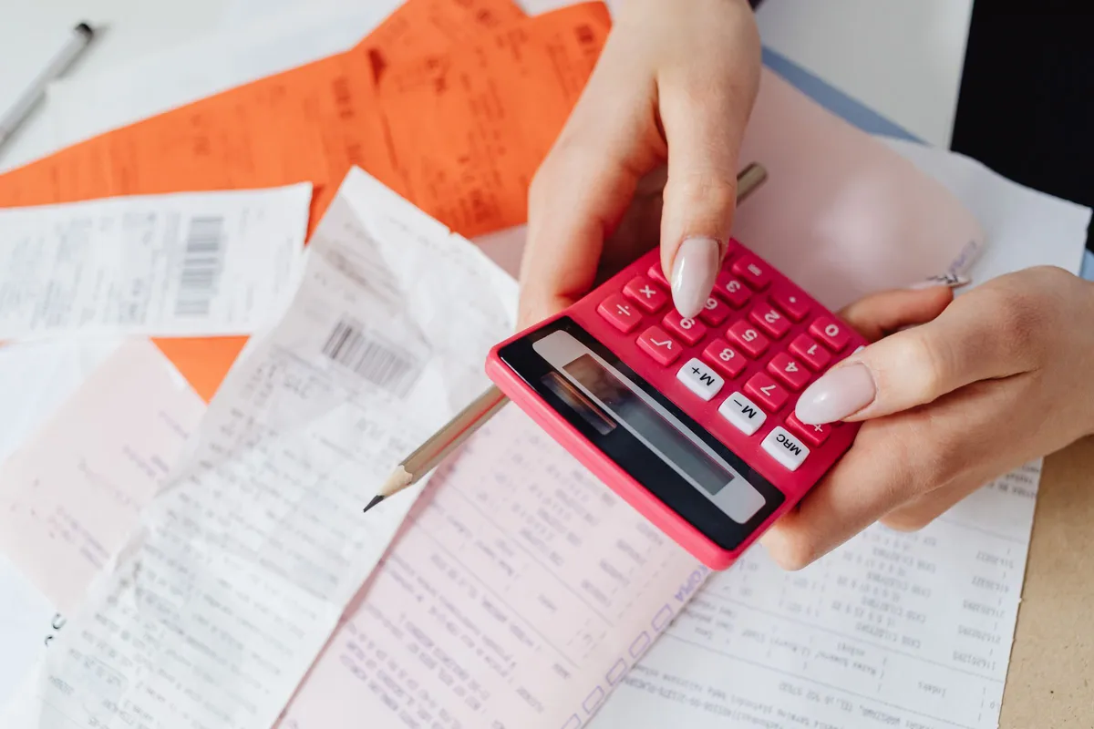 Hands tap a pink calculator over a spread of receipts and printed bills on a desk.