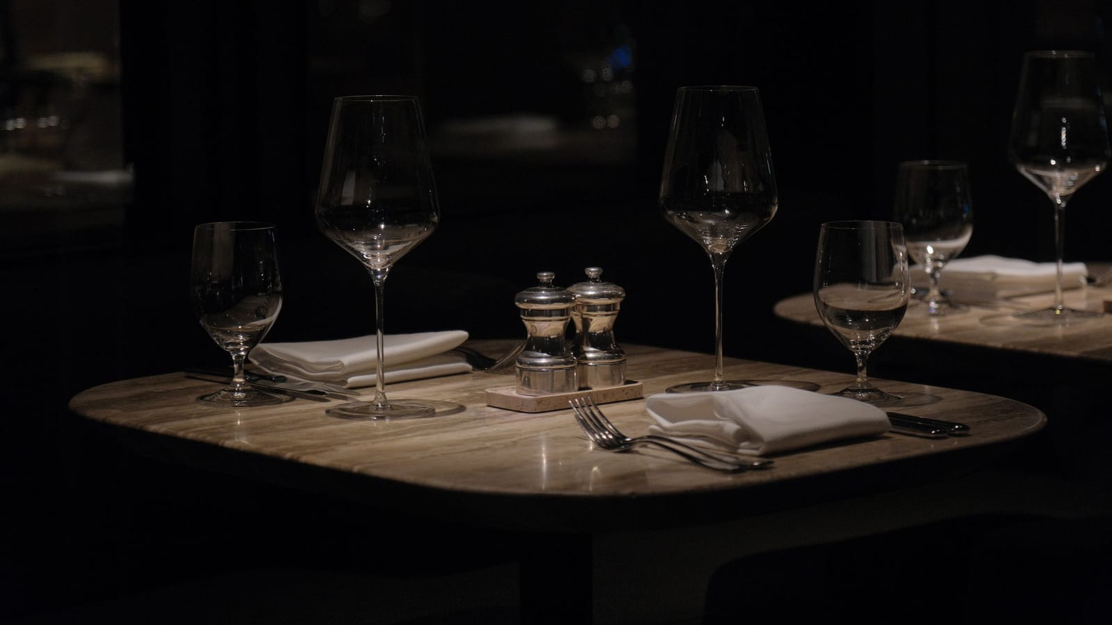 An empty restaurant table in a dark room, set with wine glasses, folded napkins and salt and pepper shakers.