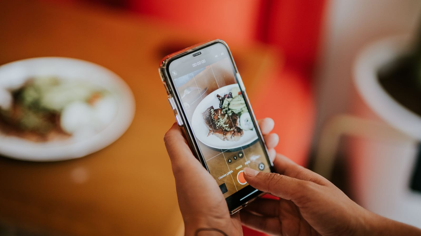 An influencer’s hands hold a phone to film a plated dish at a restaurant table.