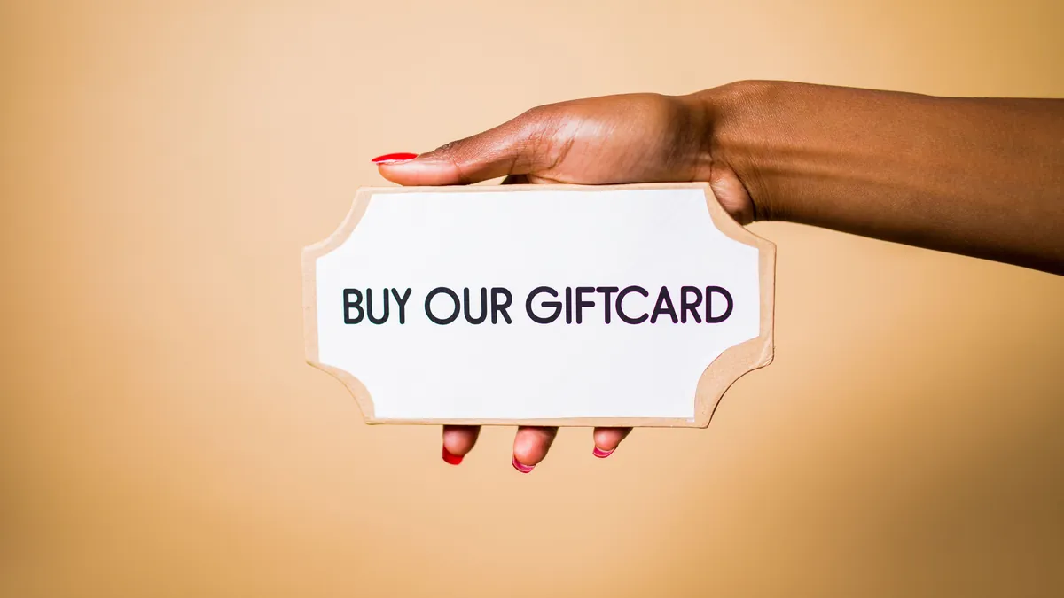 A hand holds up a white sign that reads “Buy our giftcard” against a plain peach background.