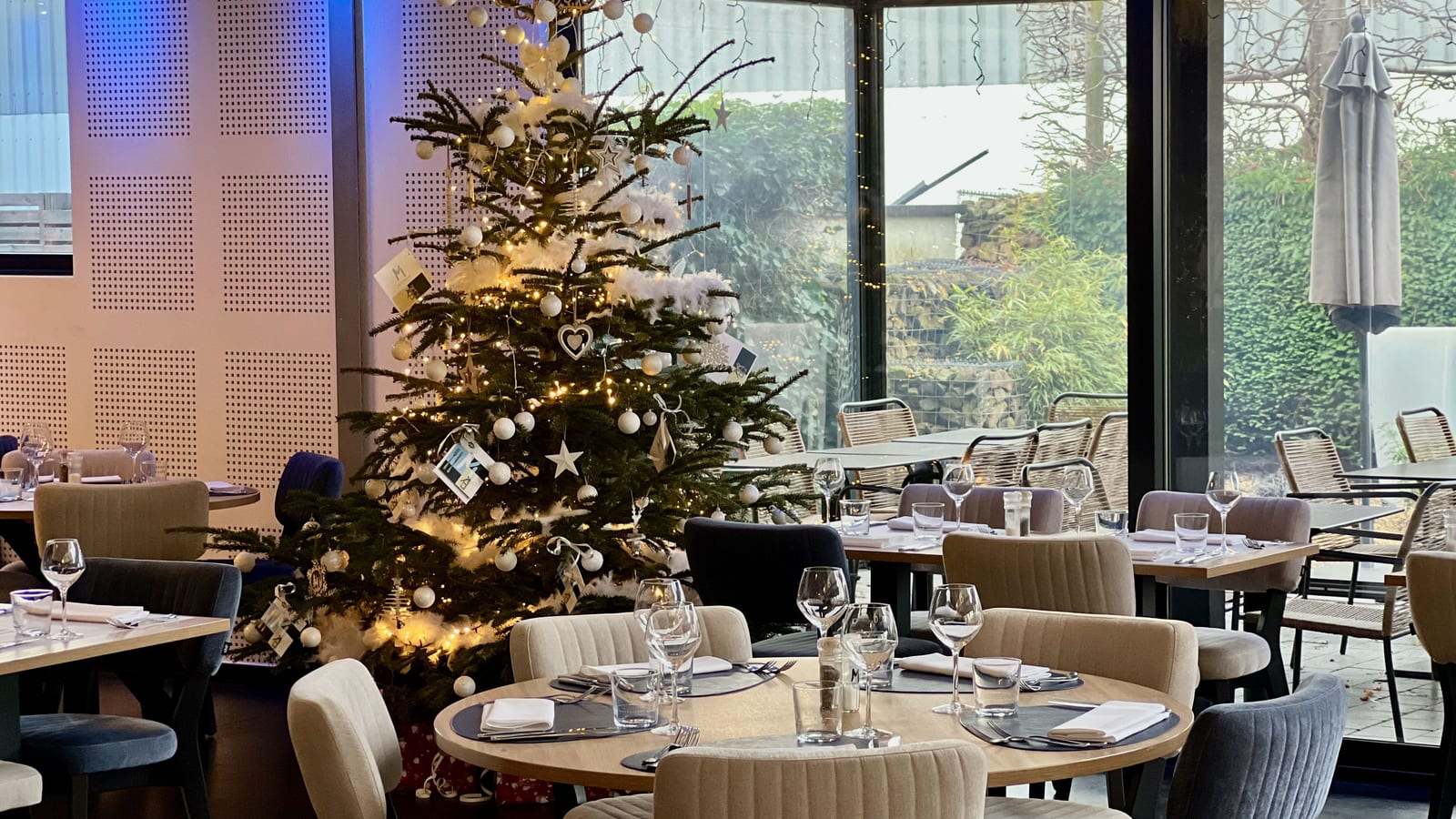 A restaurant dining room decorated for the holidays, with a lit Christmas tree beside set tables and wine glasses.