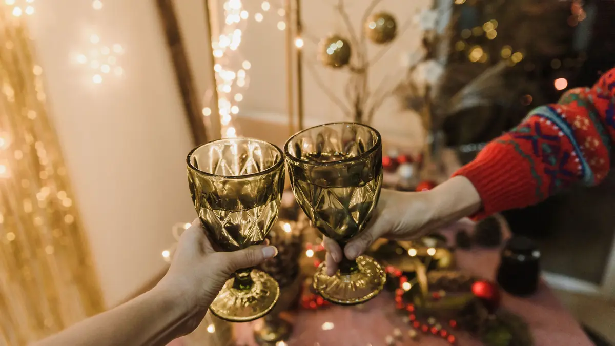 Two hands clink green glass goblets over a table of pine cones and red berries, with string lights behind.