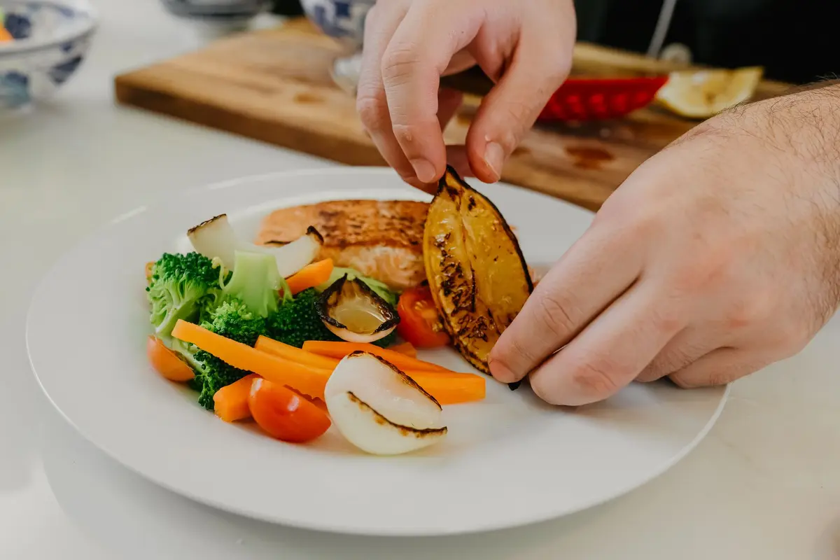 A cook’s hands set a grilled lemon slice on a white plate of salmon, broccoli, carrots and tomatoes, with a wooden board behind.