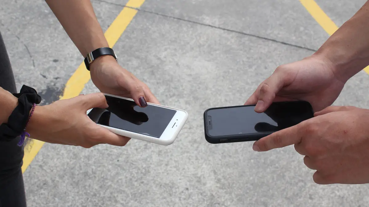 Two people hold their smartphones out toward each other over a paved lot.